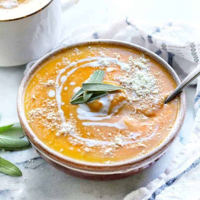 A bowl of butternut squash soup is shown on a white counter with coconut milk drizzled on top with a spoon and also fresh sage.