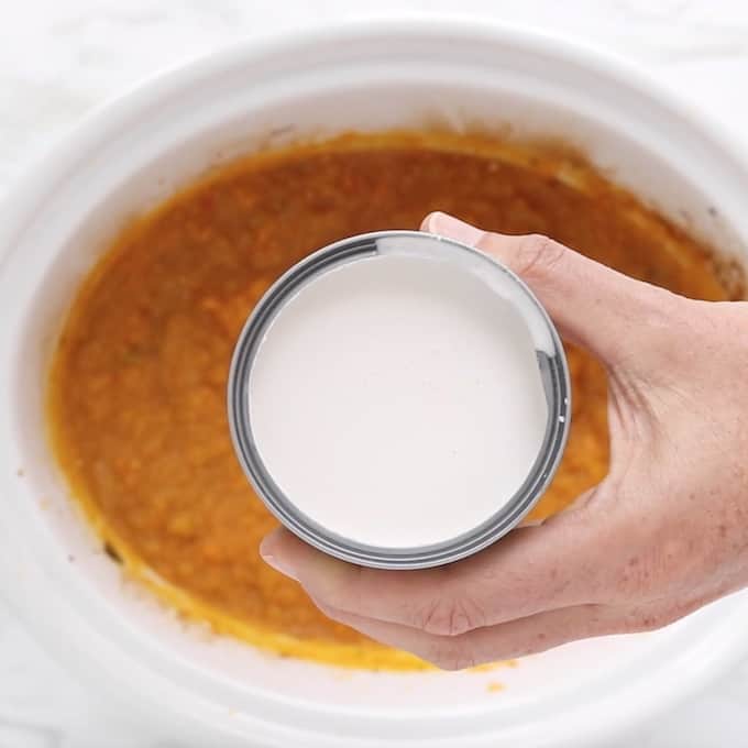 An opened can of coconut milk is held in front of the camera on top of pureed butternut squash soup in a crockpot.