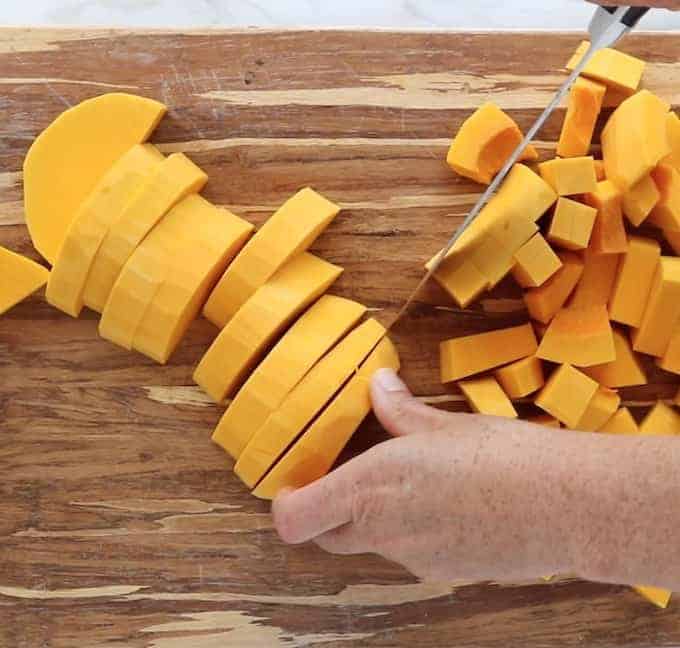A hand is slicing raw butternut squash into small slices on a cutting board.