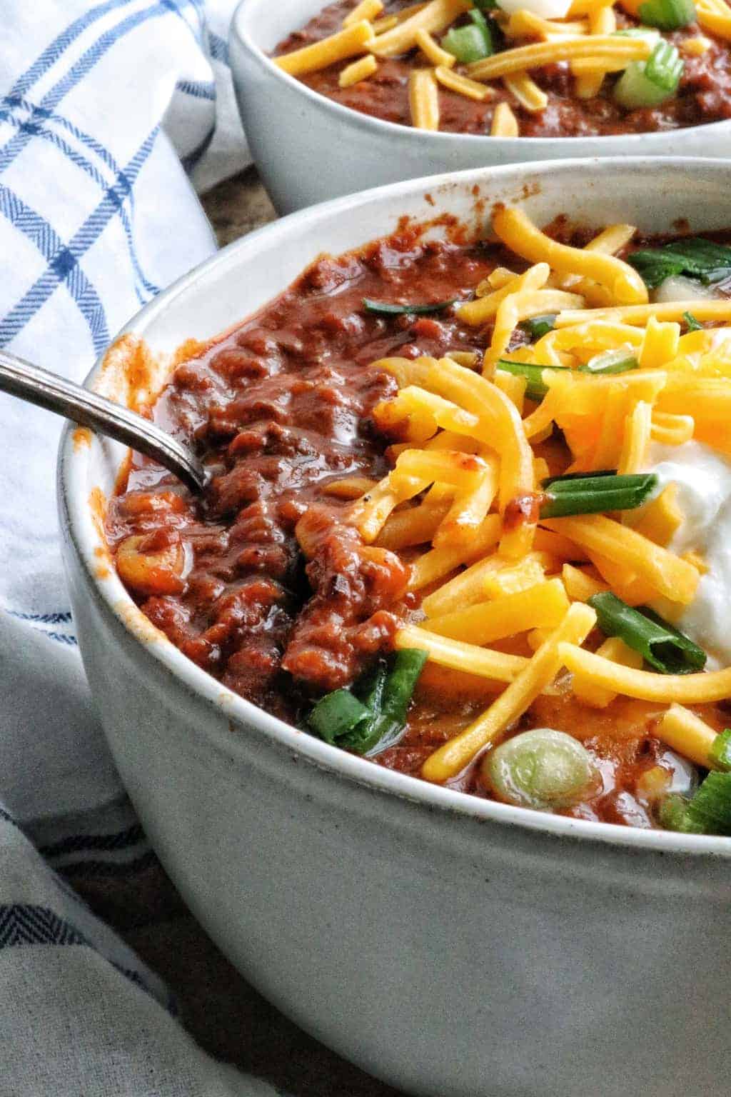 A side view of chili shown up close in a white bowl topped with shredded cheese and green onion and a spoon.