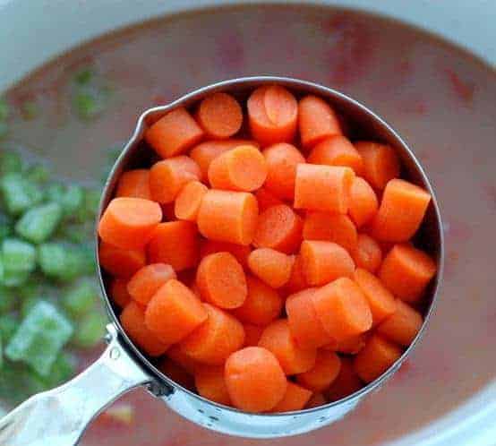 A measuring cup is holding chopped carrots up to the camera with crockpot with broth in the background.