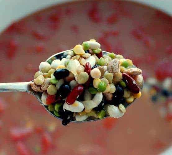 Dry bean are held on a spoon in front of the camera with the minestrone soup in the crockpot below.
