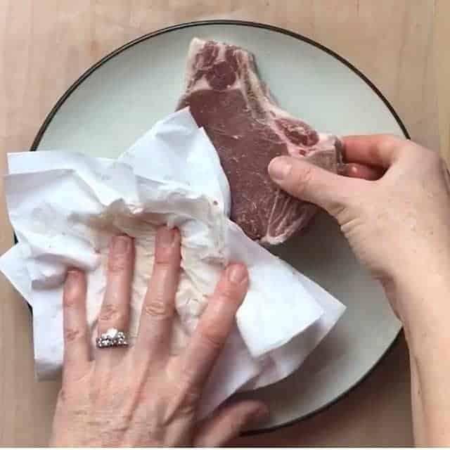 A hand is shown patting dry 2 pork chops with a paper towel. 