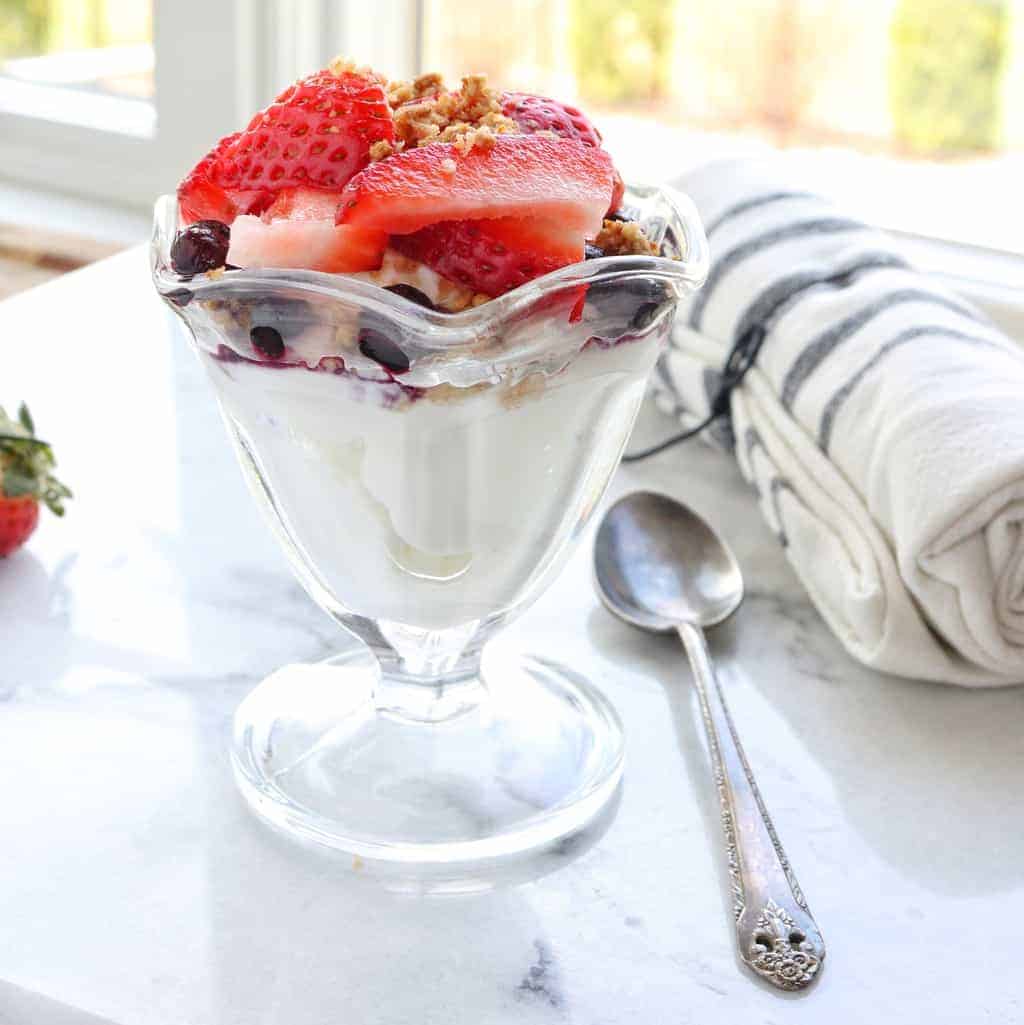 A yogurt parfait in a clear ice cream dish with slice strawberries and blueberries on top with a spoon in a bright window.