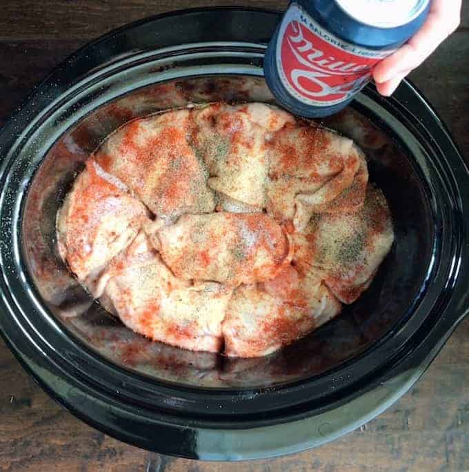 chicken thighs shown seasoned in a black crockpot with a beer being added.