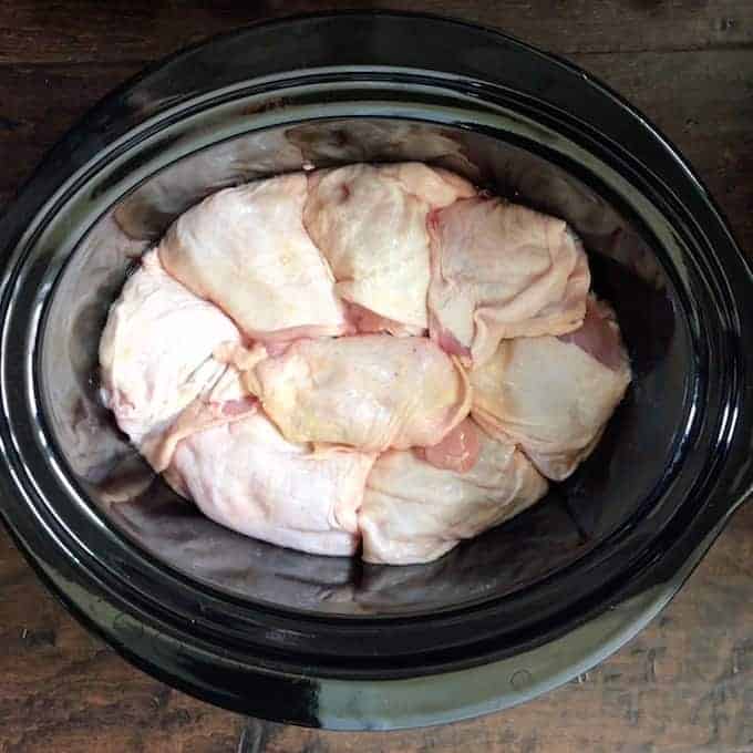 fresh or frozen chicken thighs are on bottom of a black slow cooker pot. 