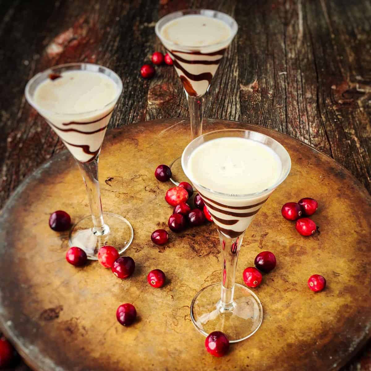 3 stemmed glasses with baileys Irish cream with chocolate swirled on the glass and cranberry on the wooden table below.