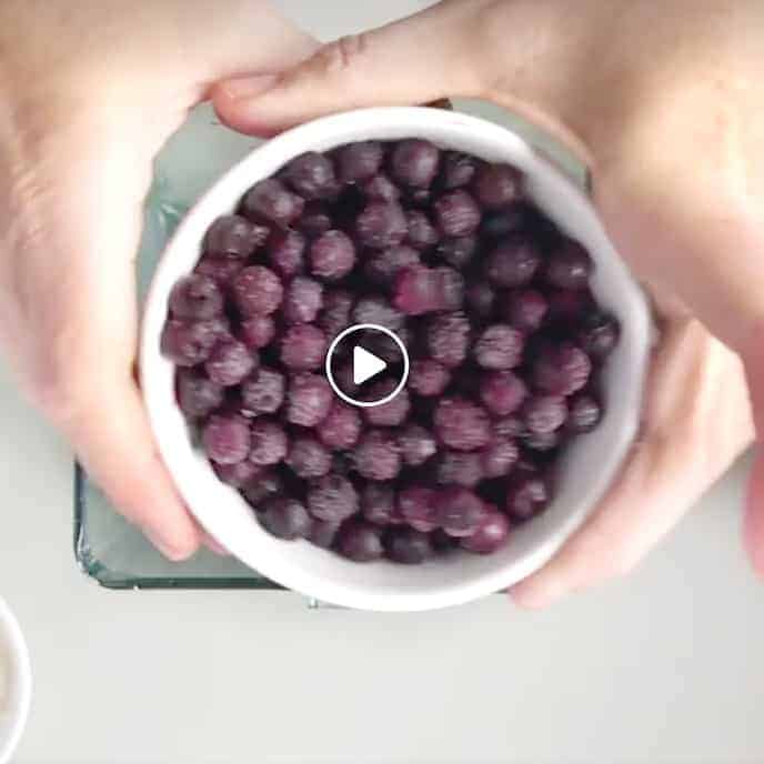 a small white bowl of frozen blueberries held in two hands above a blender on a white surface