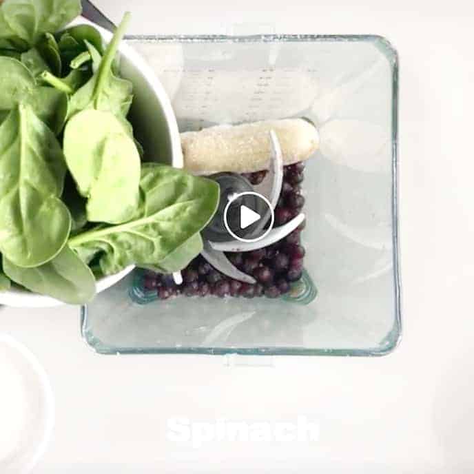 Spinach being added to a blender with frozen banana and blueberries.