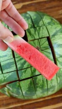 A half of a watermelon cut into watermelon sticks with one being held in front of the camera.