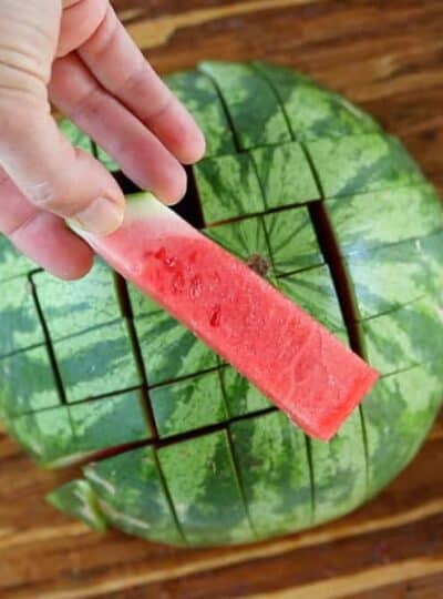A half of a watermelon cut into watermelon sticks with one being held in front of the camera.