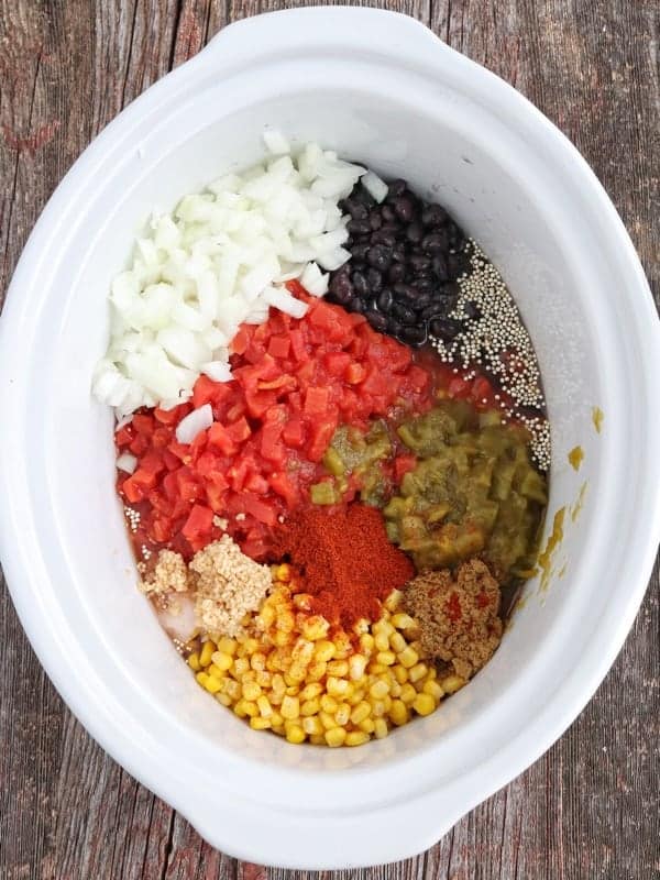 a white crockpot filled with onions, tomatoes, black beans, corn, green chilies, quinoa, broth, and spices before cooking.