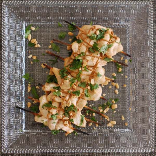 chicken satay on wooden skewers on a serving tray topped with crushed peanuts and fresh herbs.