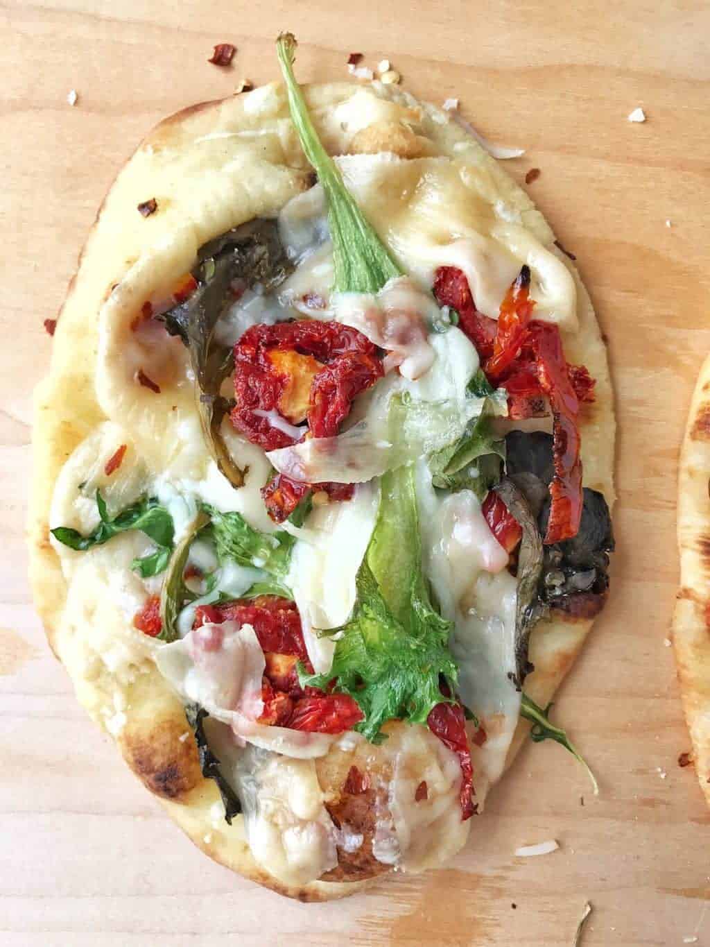 a naan pizza shown up close with parmesan cheese, garlic, leafy greens and sun dried tomatoes.