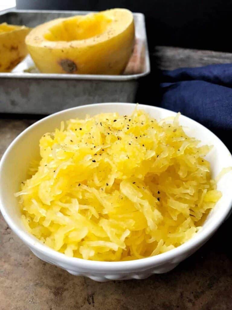 Yellow spaghetti squash strands are cooked and in a white bowl next to a cooked spaghetti squash in the shell.
