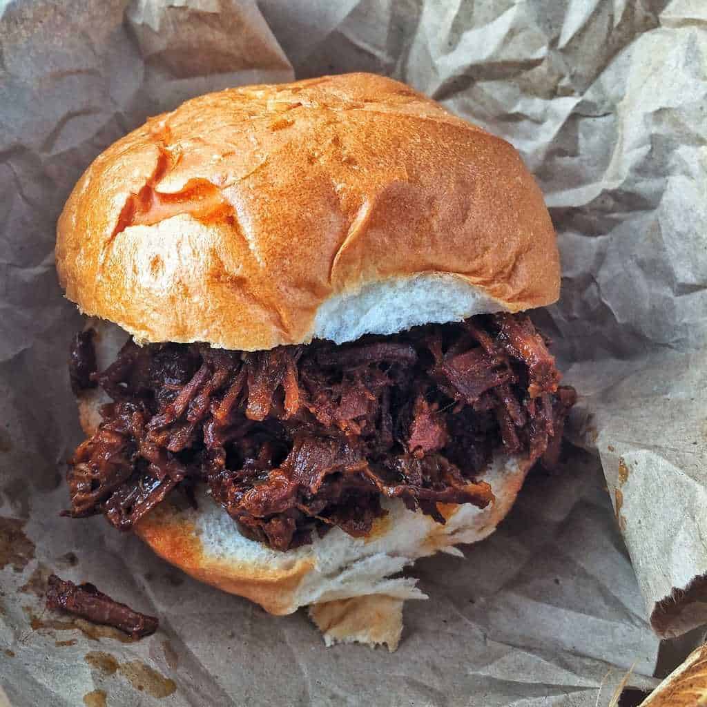 Pulled pork shown in a bun sitting on a plate lined with brown paper.