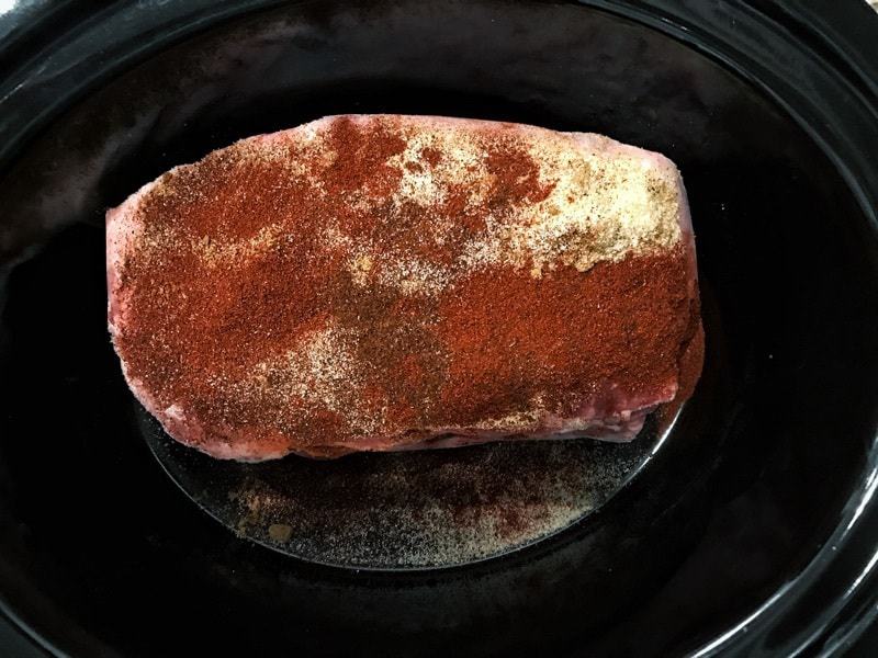 A raw pork roast with spices in a black slow cooker.