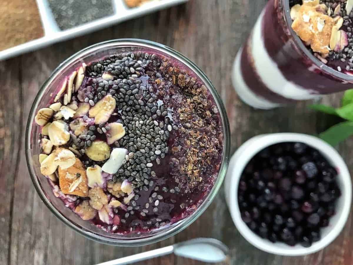 A blueberry smoothie is shown top down with granola, chia seeds, and flax seeds next to a bowl of blueberries and another smoothie.