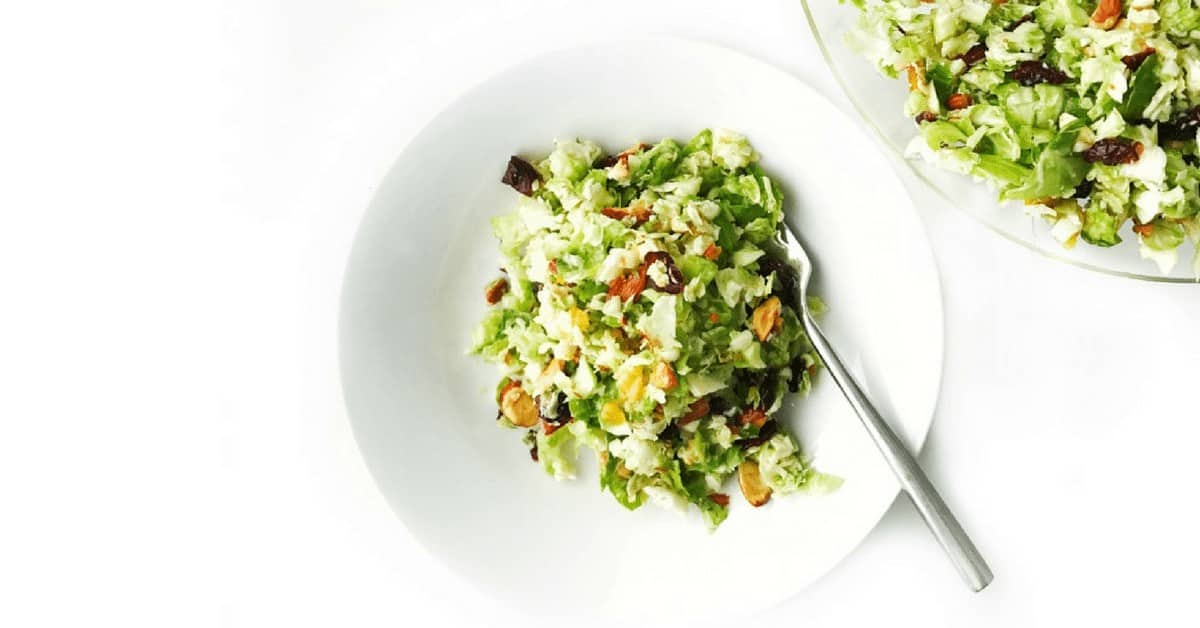 Shredded brussels sprout salad shown with almonds and dried cranberries in a white bowl with a fork on a white surface. 