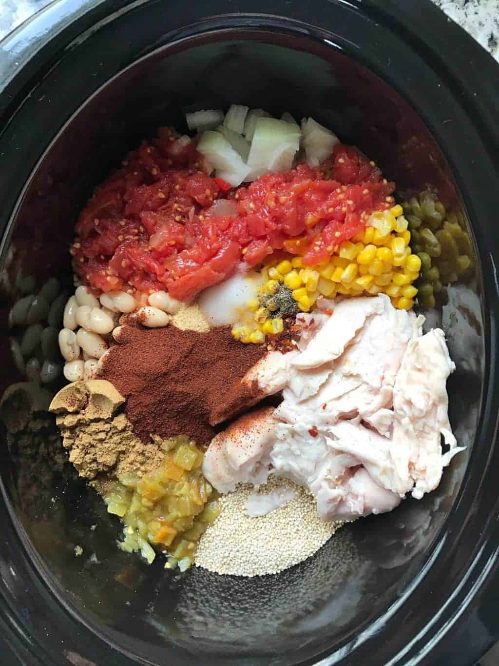 A black crockpot with frozen chicken, tomatoes, corn, spices, beans, dry quinoa, and onions.