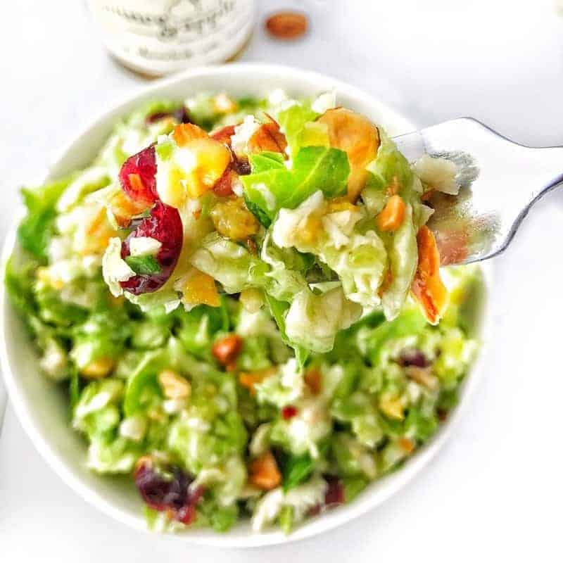 Shredded brussels sprout salad shown with almonds and dried cranberries in a white bowl with a fork holding a bite up close.