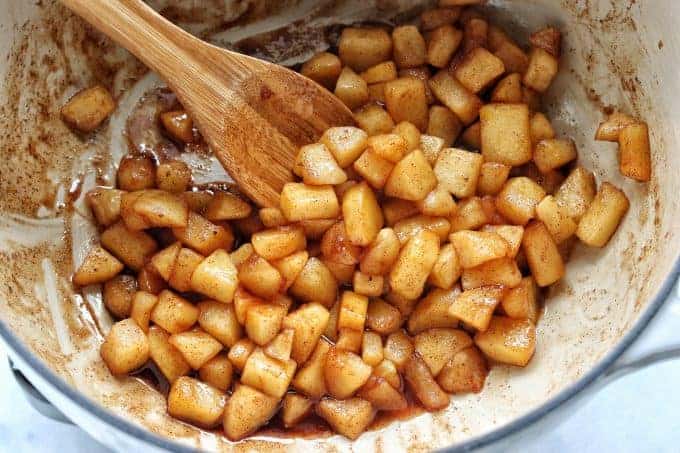 diced apples shown cooked in a white pot with a wooden spoon stirring the baked apples coated with cinnamon and maple syrup sauce. 