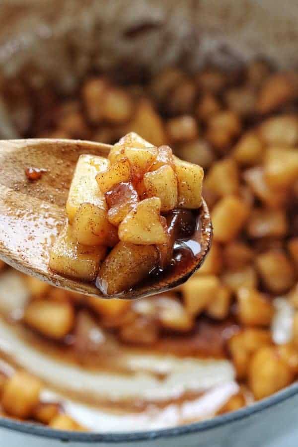 Close up of a baked apples shown on a wooden spoon with more baked apples in a white pot in the background.