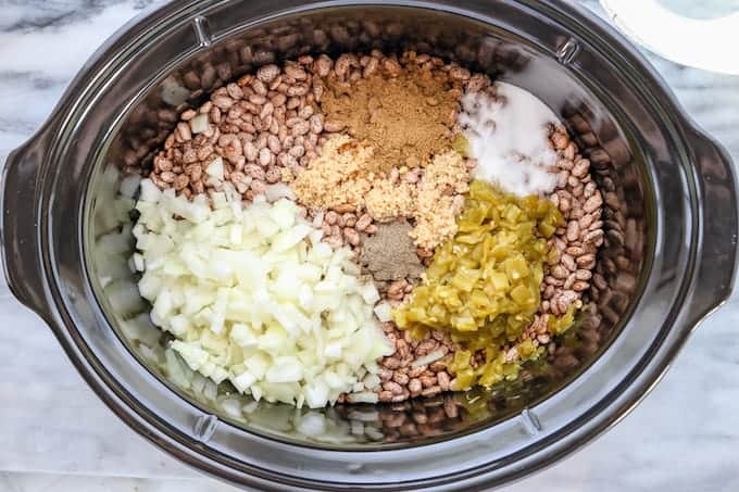 diced onion, dry pinto beans, and spices in a black crockpot on a white surface.