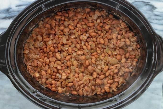 cooked beans and onions before mashing in a black crockpot on a white surface.