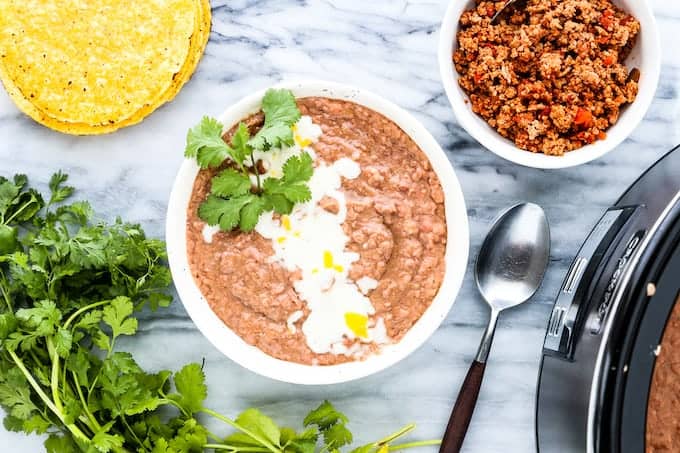 a white bowl with refried beans, melted cheese and cilantro next to the crockpot with beans, tortilla shells and a bowl of taco meat. 