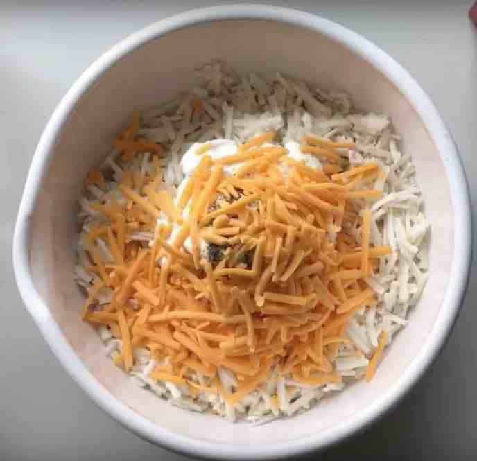 A large white bowl is filled with shredded hash browns, greek yogurt, cream cheese, spices, and shredded cheddar cheese. 
