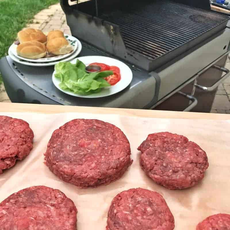 raw formed cheese stuffed burgers are on a tray by an out door grill. 