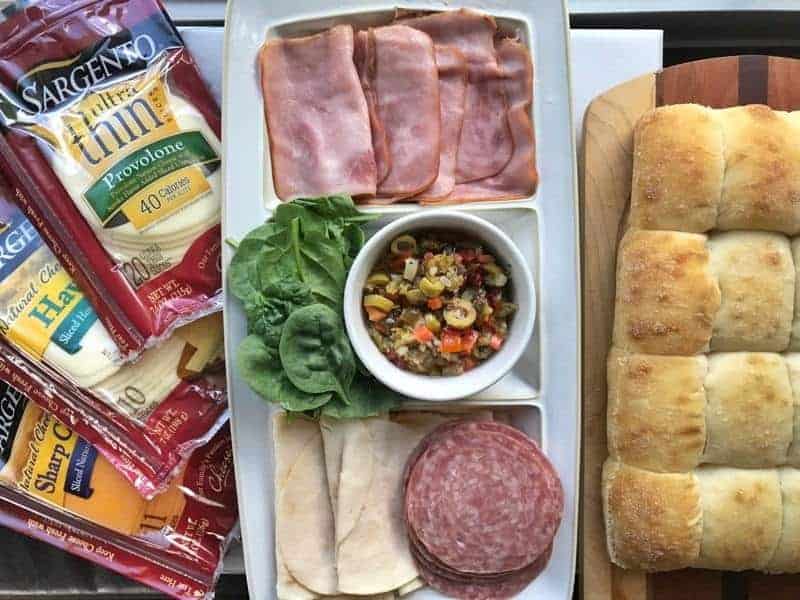 packaged cheese, ham, turkey, salami, olive blend, and rolls shown on a cutting board.