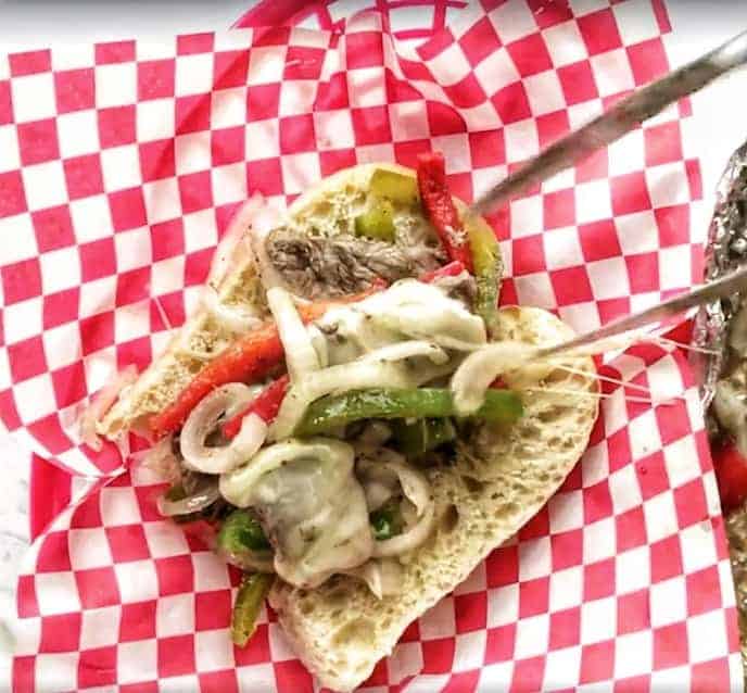 Philly Cheese Steak sandwich showing a roll being piled with beef, peppers, onions and cheese in a basket with red and white checkered paper.