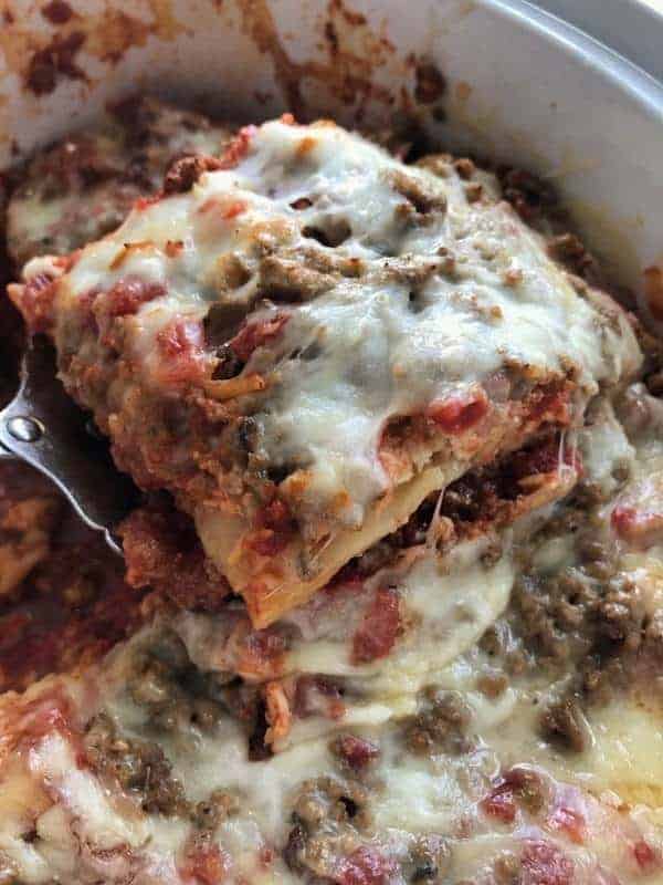 slow cooker lasagna in a white slow cooker with a spatula scooping up a cheesy slice. 