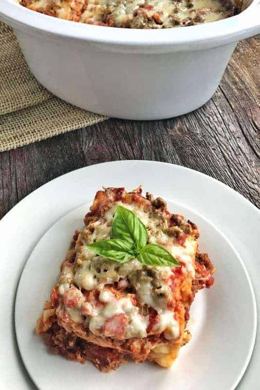 slow cooker lasagna on a white plate with the white slow cooker behind with the rest of the lasagna inside. 