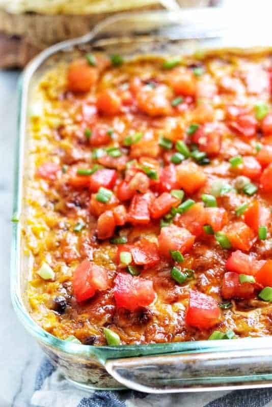 Seven layer bean dip shown baked with tomatoes and green onions on top in a glass casserole pan. 