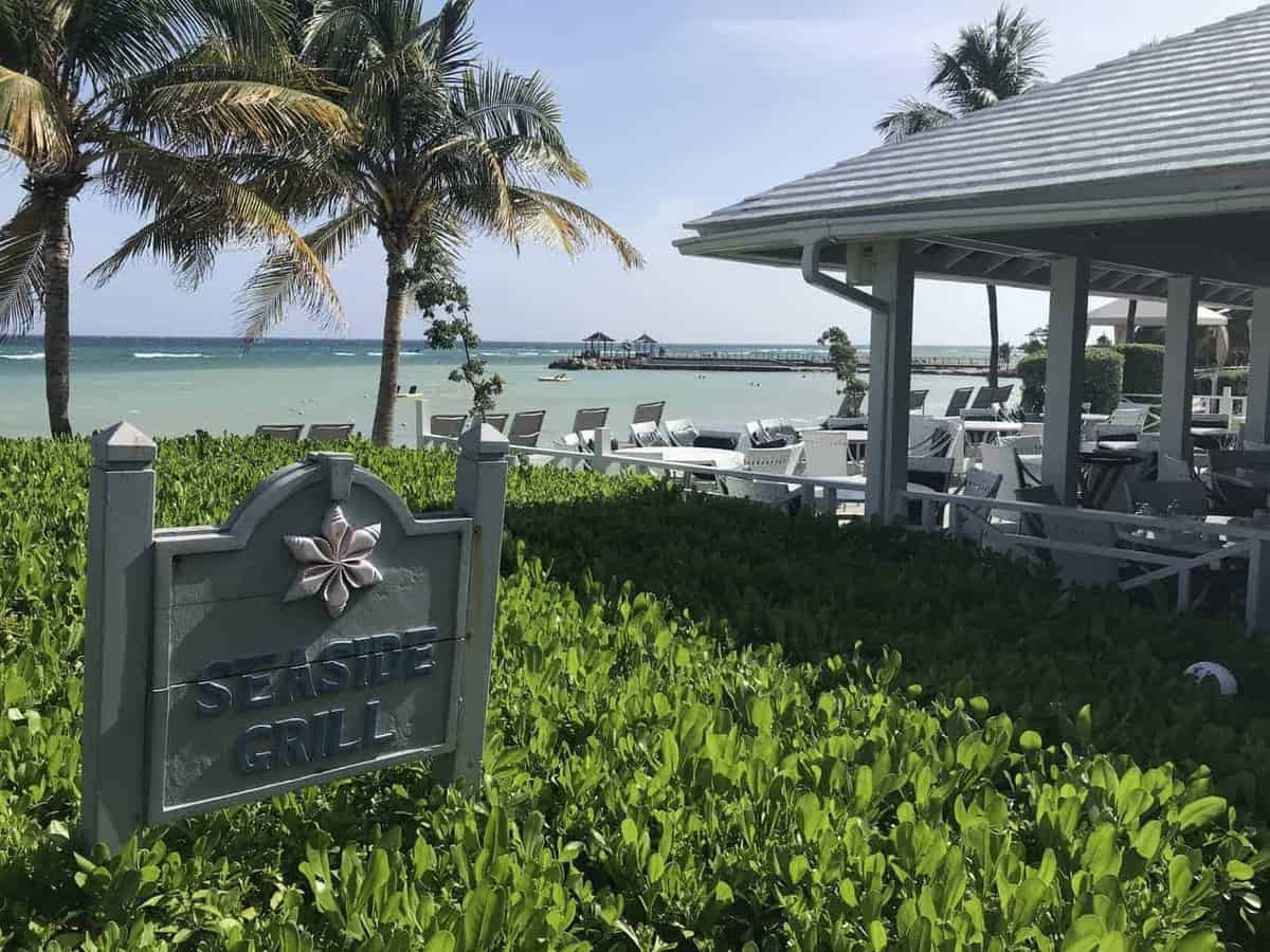 The seaside grill at the Hilton Rose Hall Jamaica set right in front of the beach with chairs and palm trees. 