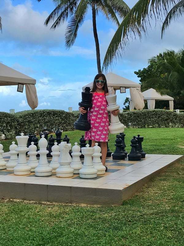 Little girl standing a large outdoor chess set holding pieces under her arms in the palm trees.