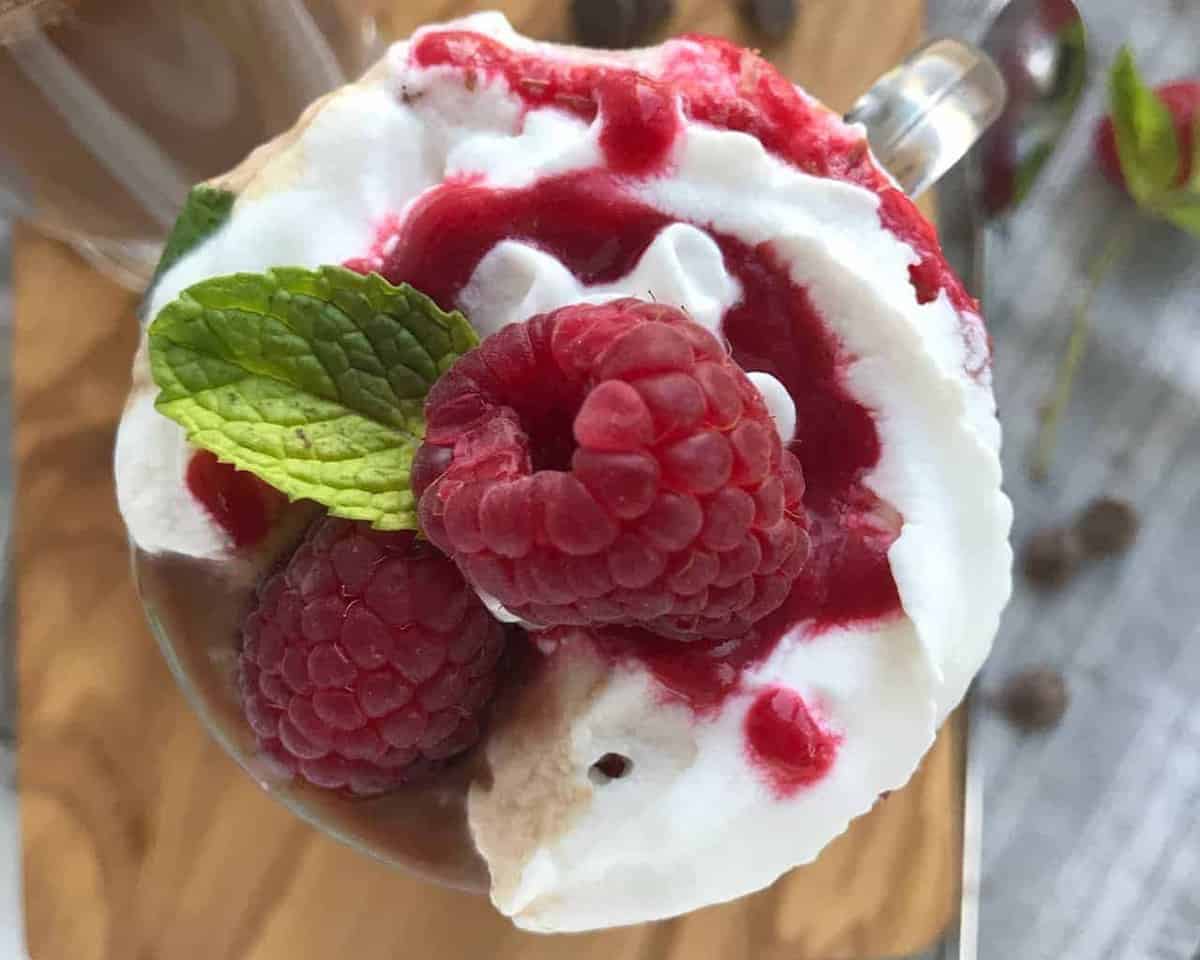 a clear mug of hot chocolate with whipped cream and a raspberry on top
