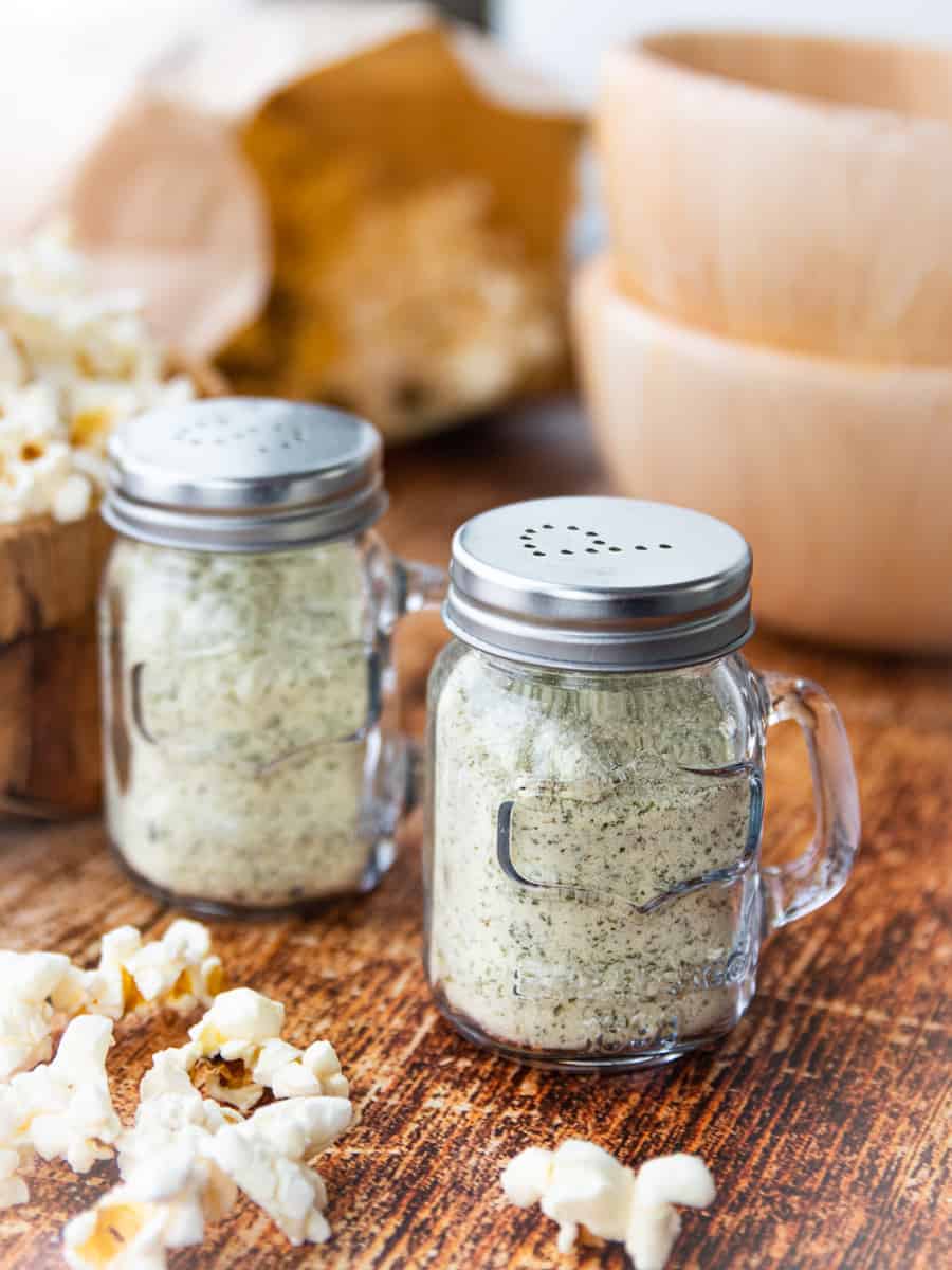 Ranch popcorn seasoning is shown in 2 glass containers on a wooden surface with 2 wooden bowls and popped popcorn in the background. 