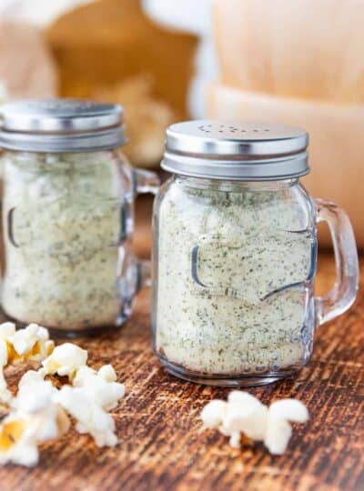 two shakers of ranch popcorn seasoning on a table with popcorn.