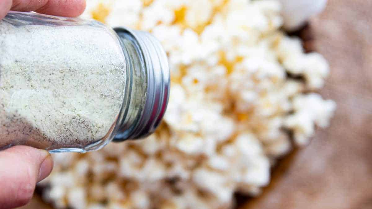 Ranch popcorn seasoning in a shaker being sprinkled over a bowl of popped popcorn on a wooden surface.