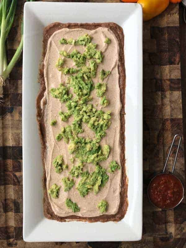 Refried bean dip recipe shown with refried beans, Greek Yogurt mixture and avocado spread on a white serving tray.