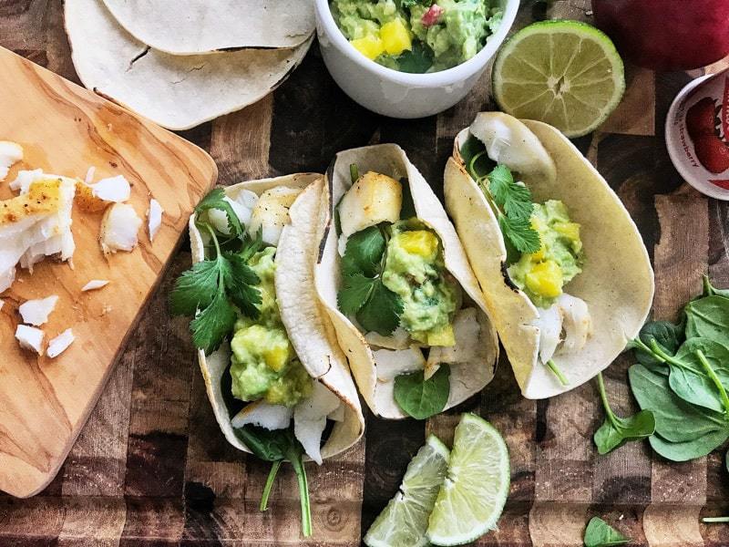 3 Baked Cod Fish tacos with mango guacamole on a wooden surface with lime and spinach. 