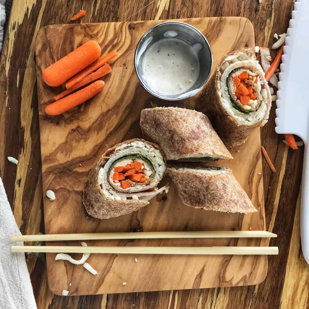 A turkey roll up is sliced on a wooden try with carrots and ranch. Inside the wrap is turkey, spinach, and carrots.