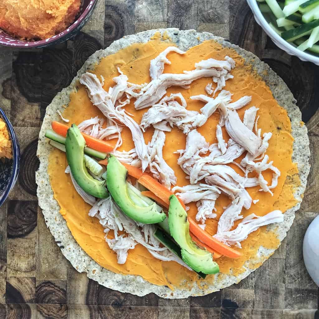 A flour tortilla is spread with spread cheese layered with chicken and on one side carrots and avocado slices.