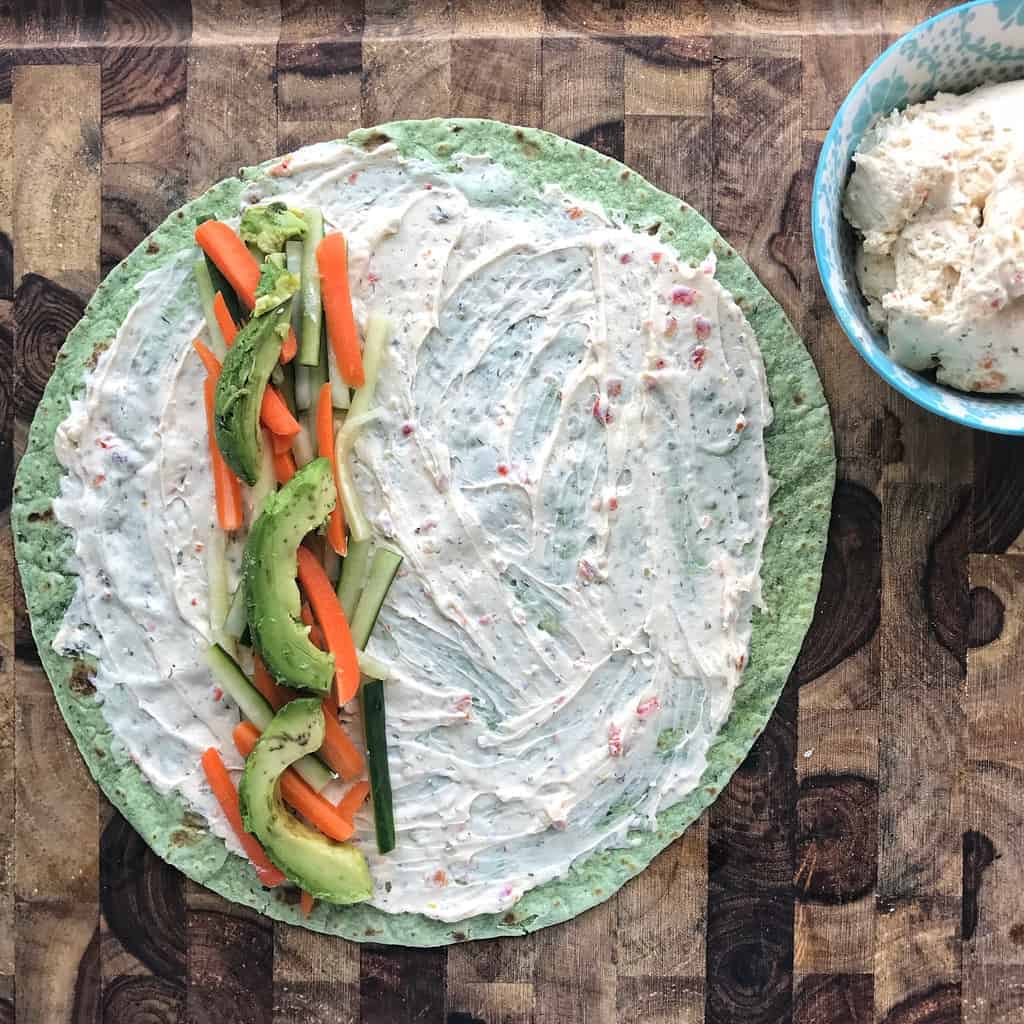 A spinach flour tortilla is spread with cheese spread and on one side is cucumber, carrots, and avocado slices. 