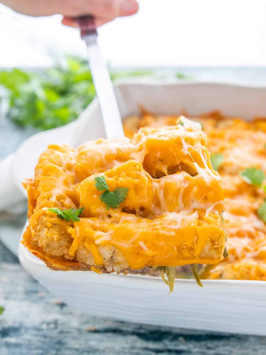 healthy tater tot casserole shown in a white bowl topped with cheese with a slice being removed from the pan.