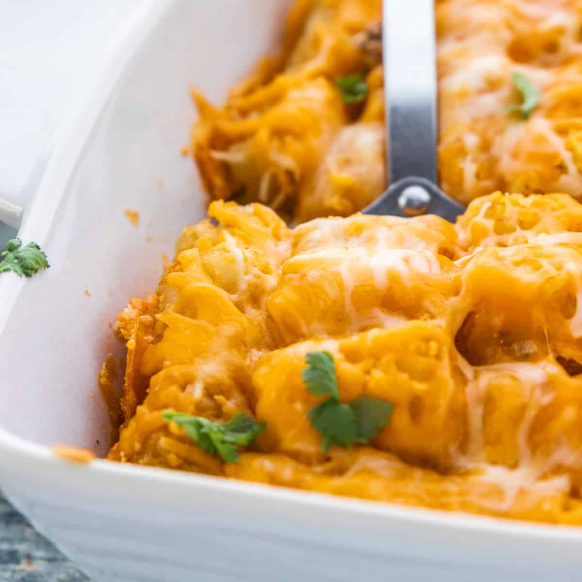 tater tot casserole shown close up in a white baking dish with a spatula lifting up a slice. 